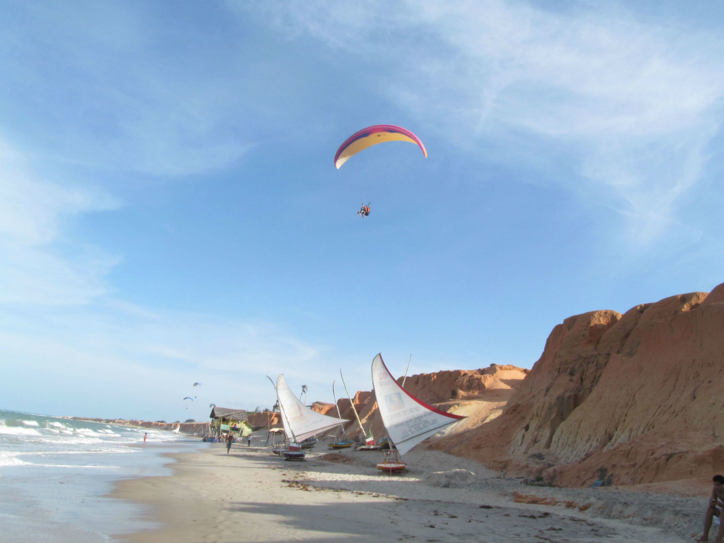A beach with several sailboats anchored near the shore, a clear sky, and rocky cliffs in the background.