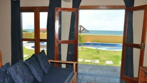 A room with a sofa and a coffee table, a large window with a view of a beach and ocean, and a door with a wooden frame. - Sirius Canoa Residencial (Aracati)