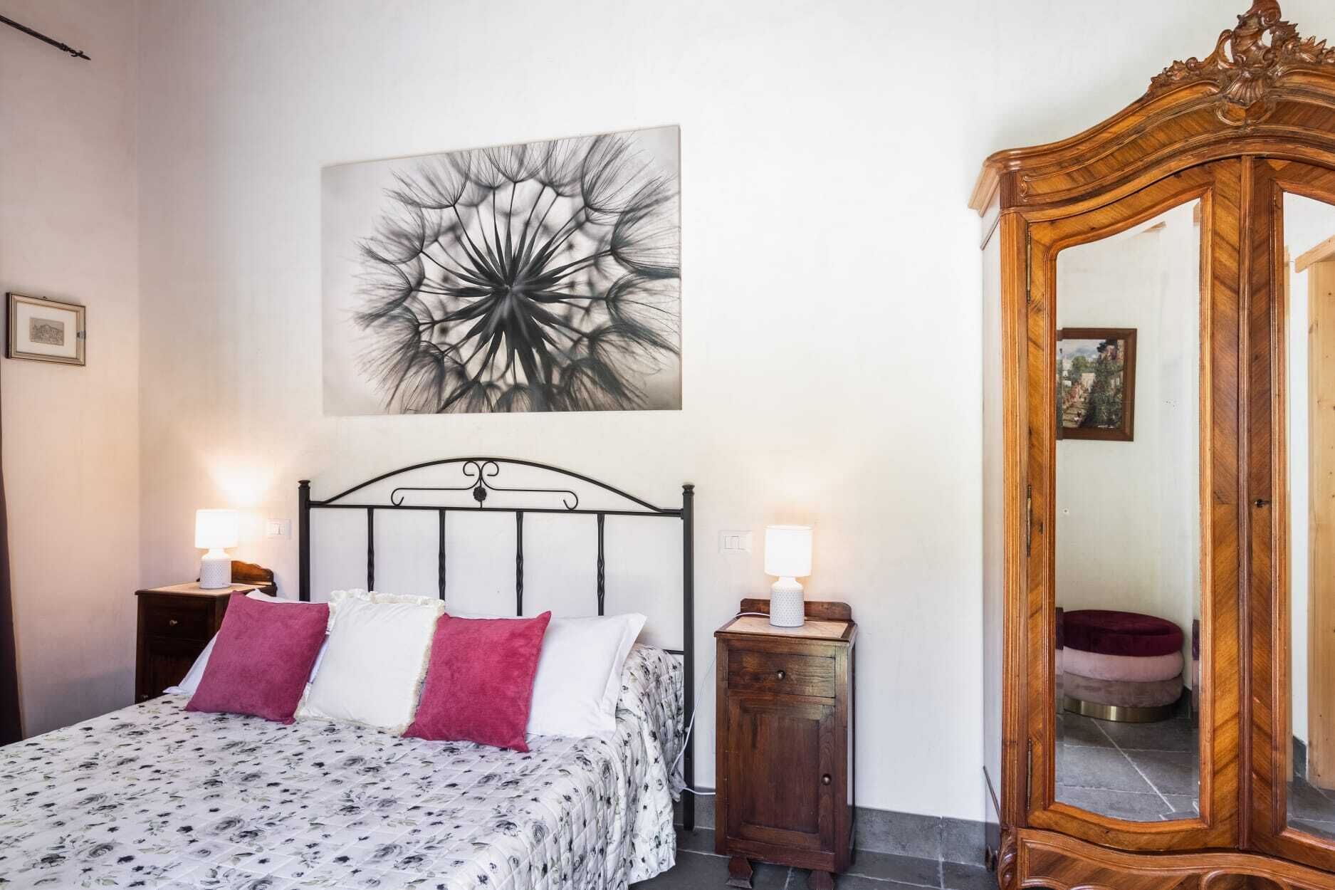 A bedroom with a bed, two bedside lamps, a wooden dresser, a large mirror, and a floral artwork on the wall.
