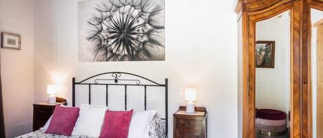A bedroom with a bed, two bedside lamps, a wooden dresser, a large mirror, and a floral artwork on the wall.