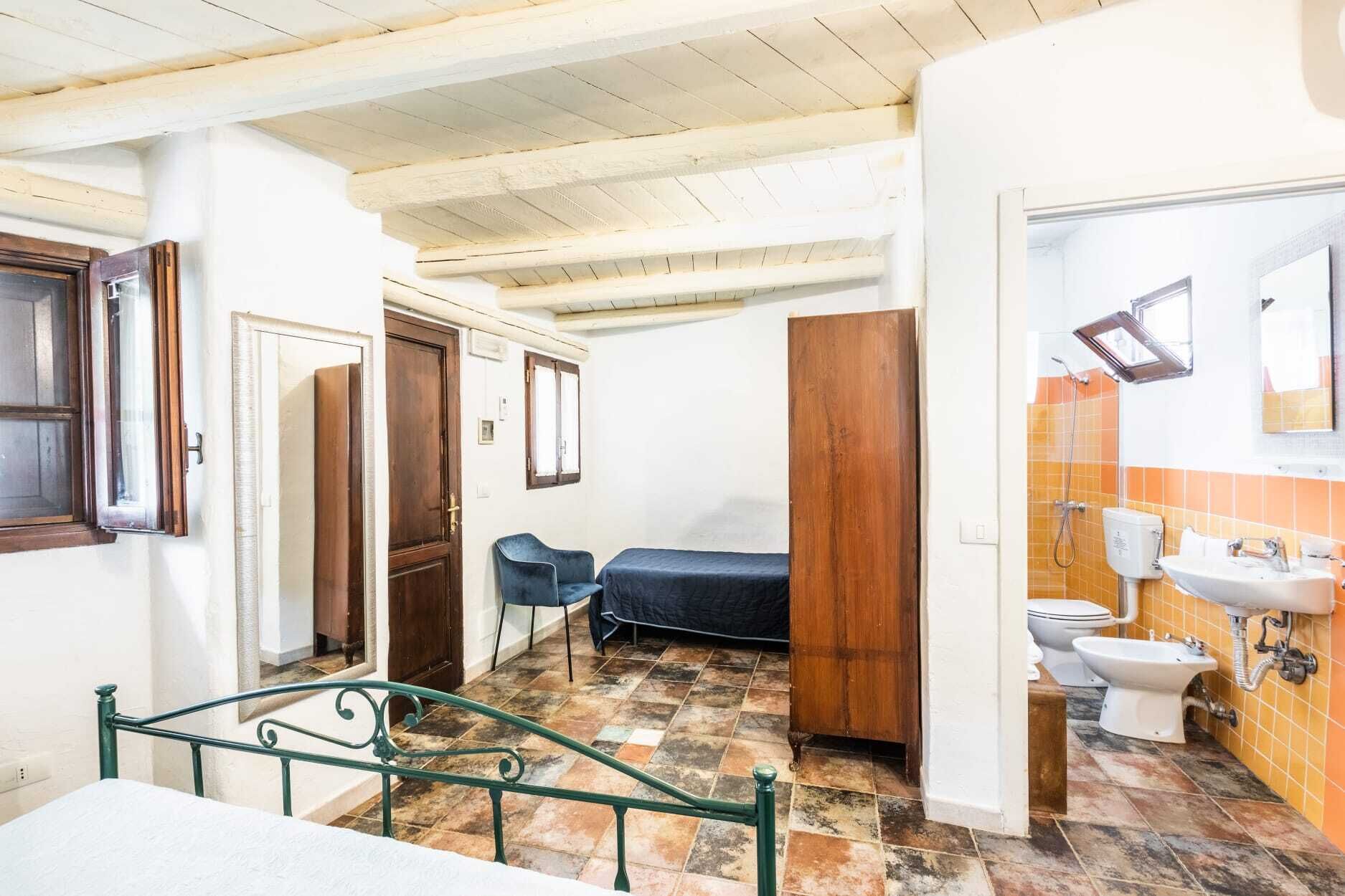 Una habitación con una cama, una mesita, una silla, un armario de madera y un baño con inodoro y lavamanos.