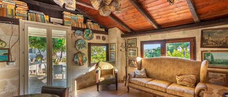 A cozy room with a wooden ceiling, a sofa, a chair, and a table. There are books and decorative items on shelves.