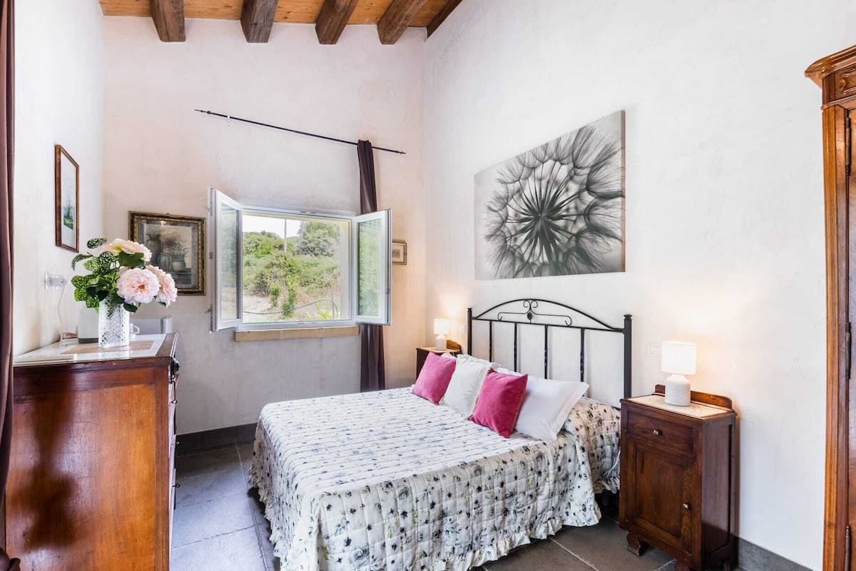 Een slaapkamer met een bed, een raam, een nachtkastje, een lamp, een fotolijst en een vaas met bloemen.