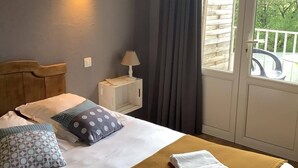 A hotel room with a bed, a nightstand, a lamp, a window with curtains, and a view of greenery outside. - Logis Hôtel La Borie (Saint-Genies)