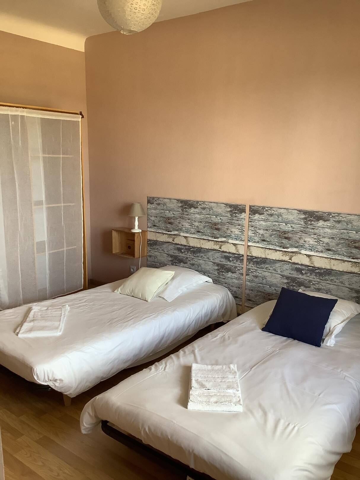 Una habitación con dos camas, cabecero de madera, una mesita de noche con lámpara y una ventana con cortina translúcida.