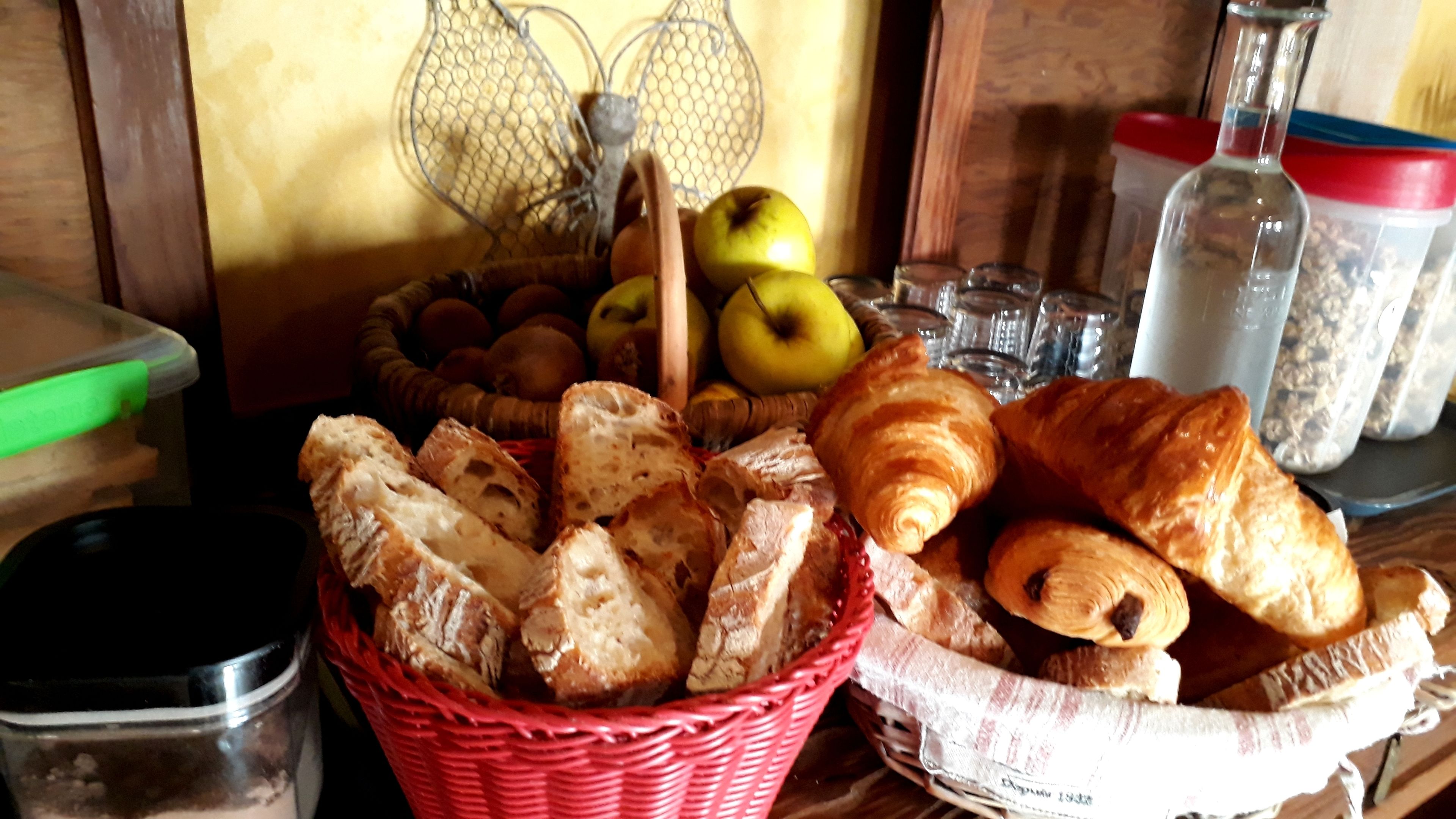 A basket of bread and pastries, a wire fruit basket with apples, and a glass jar on a wooden surface.
