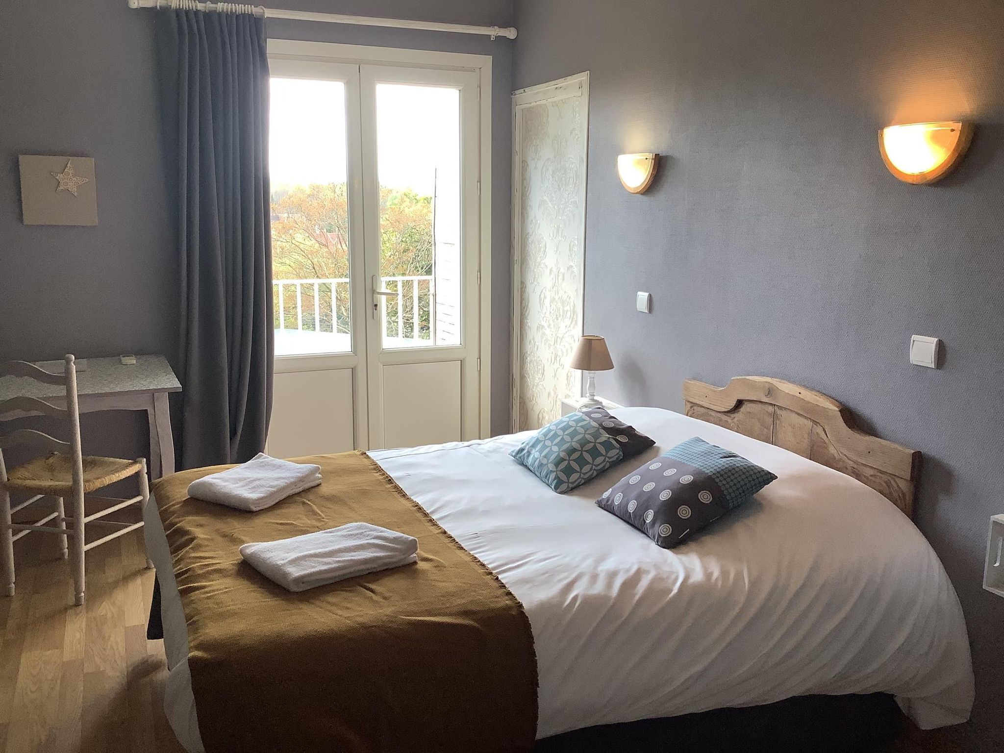 A hotel room with a bed, a wooden headboard, a nightstand with a lamp, a window with curtains, and a view of trees outside.