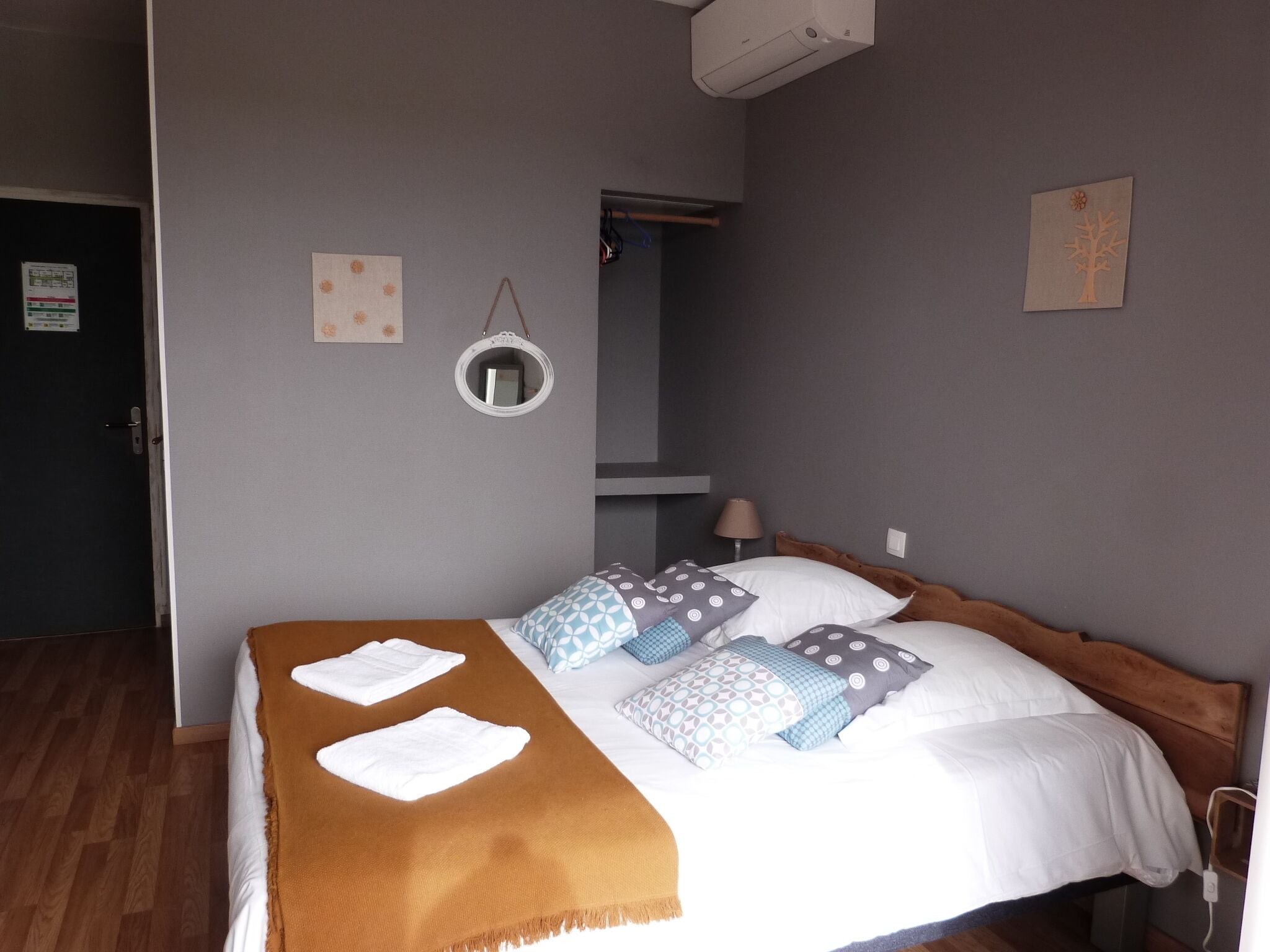 A neatly made bed with a brown blanket and patterned pillows, a round mirror on the wall, and a door to the left.