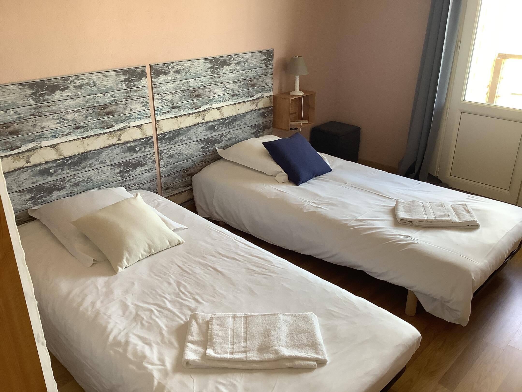 A room with two beds, each with white bedding and a folded towel. There is a wooden headboard with a distressed finish, a small table with a lamp, and a window with curtains.