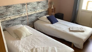 Une chambre avec deux lits, chacun recouvert d’une literie blanche et agrémenté d’une serviette pliée. On y trouve une tête de lit en bois au fini vieilli, une petite table avec une lampe et une fenêtre avec des rideaux.