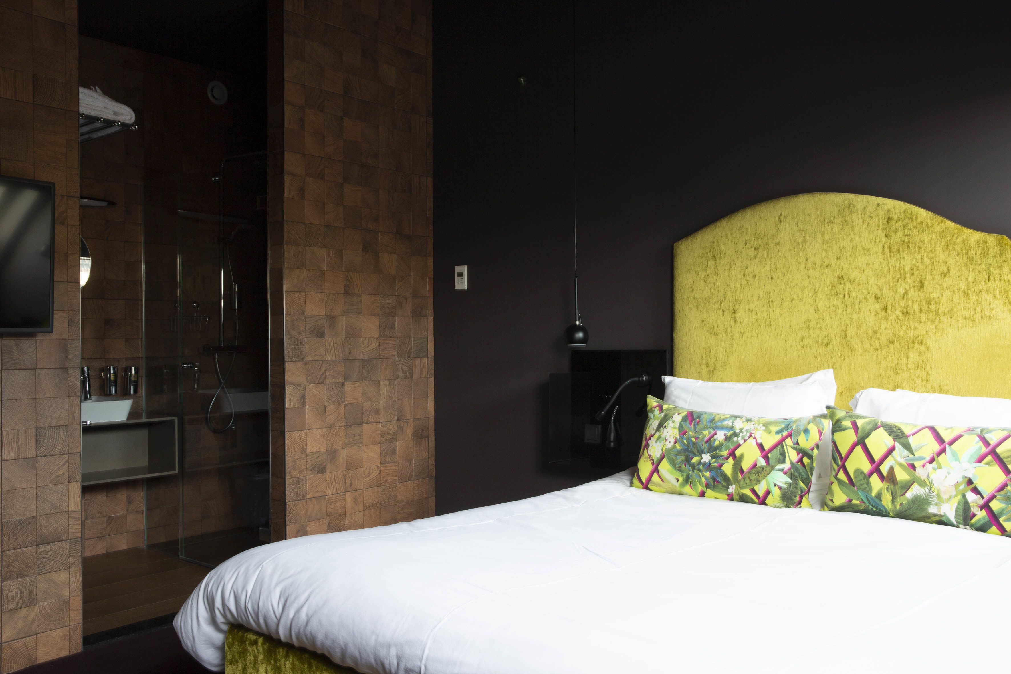 A hotel room with a bed, a TV, a bathroom with a glass shower enclosure, and a yellow headboard.