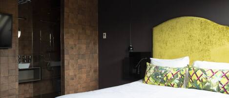 A hotel room with a bed, a TV, a bathroom with a glass shower enclosure, and a yellow headboard.