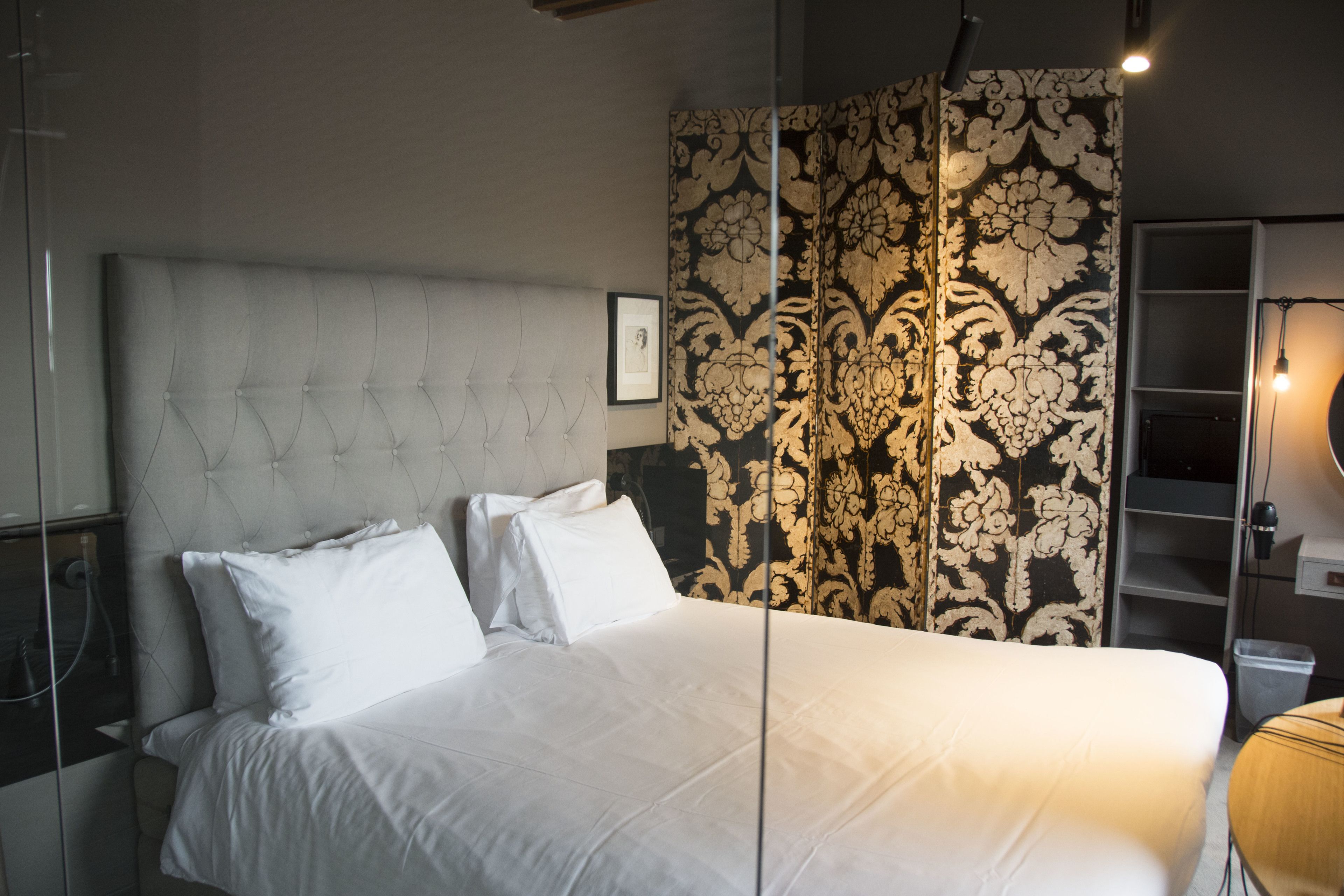 A neatly made bed with white linens and a tufted headboard, a patterned wallpaper, and a bedside lamp.
