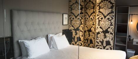 A neatly made bed with white linens and a tufted headboard, a patterned wallpaper, and a bedside lamp.