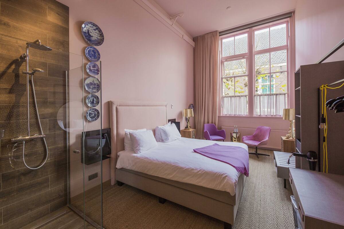 A hotel room with a glass-enclosed shower, a bed with white linens, a desk with a chair, and a window with curtains.