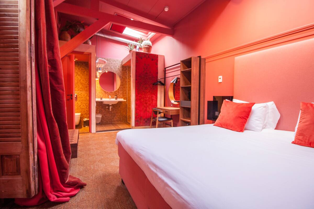 A hotel room with a bed, a desk, a chair, a mirror, a wardrobe, and a bathroom with a shower and toilet.