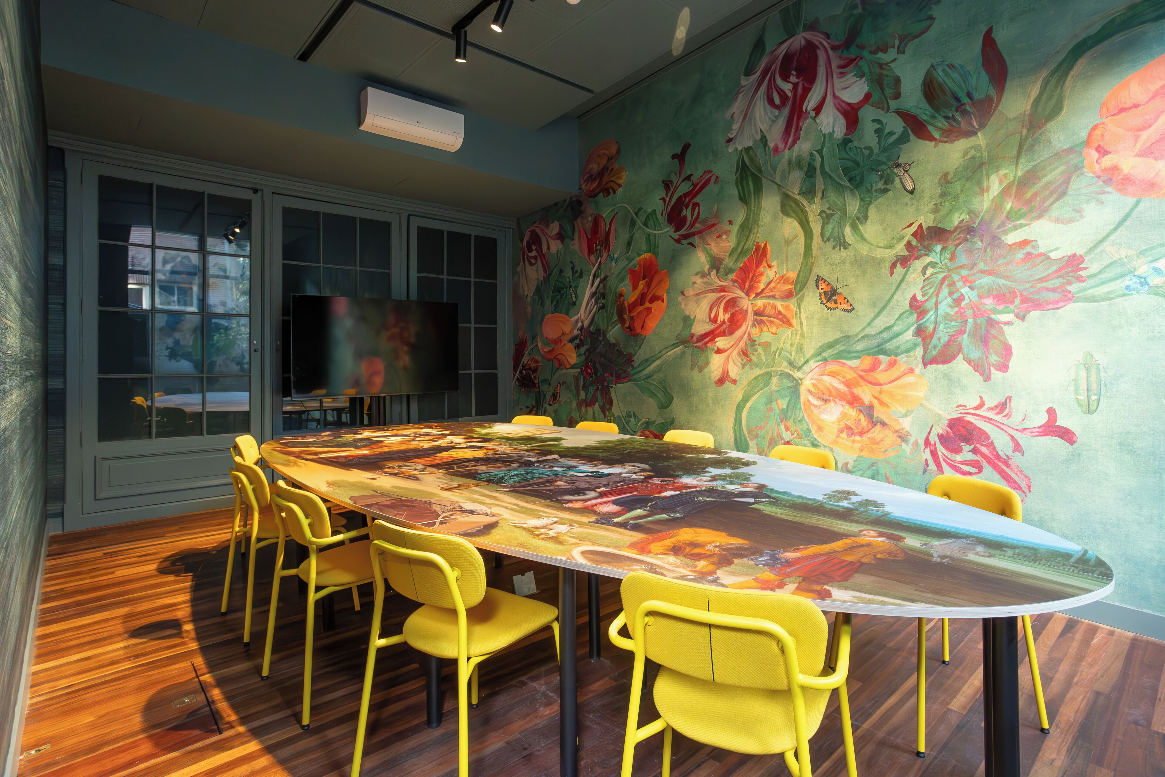 A conference room with a large oval table, yellow chairs, and a floral mural on the wall.