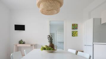 A modern dining area with a white table, chairs, a fruit bowl, a woven light fixture, a flat-screen TV, and a refrigerator.