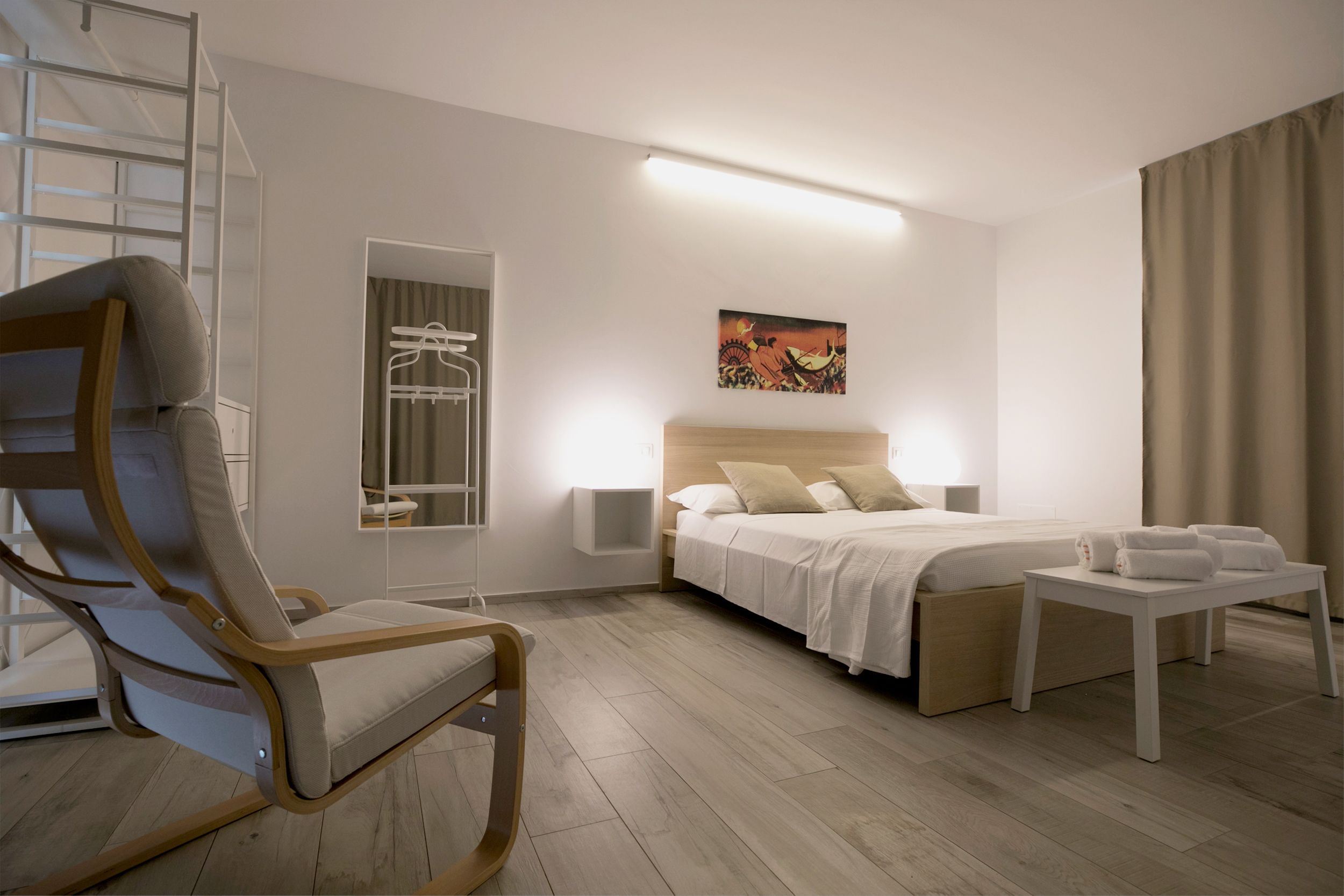 A modern hotel room with a bed, a chair, a bedside table with towels, a mirror, and a painting on the wall.
