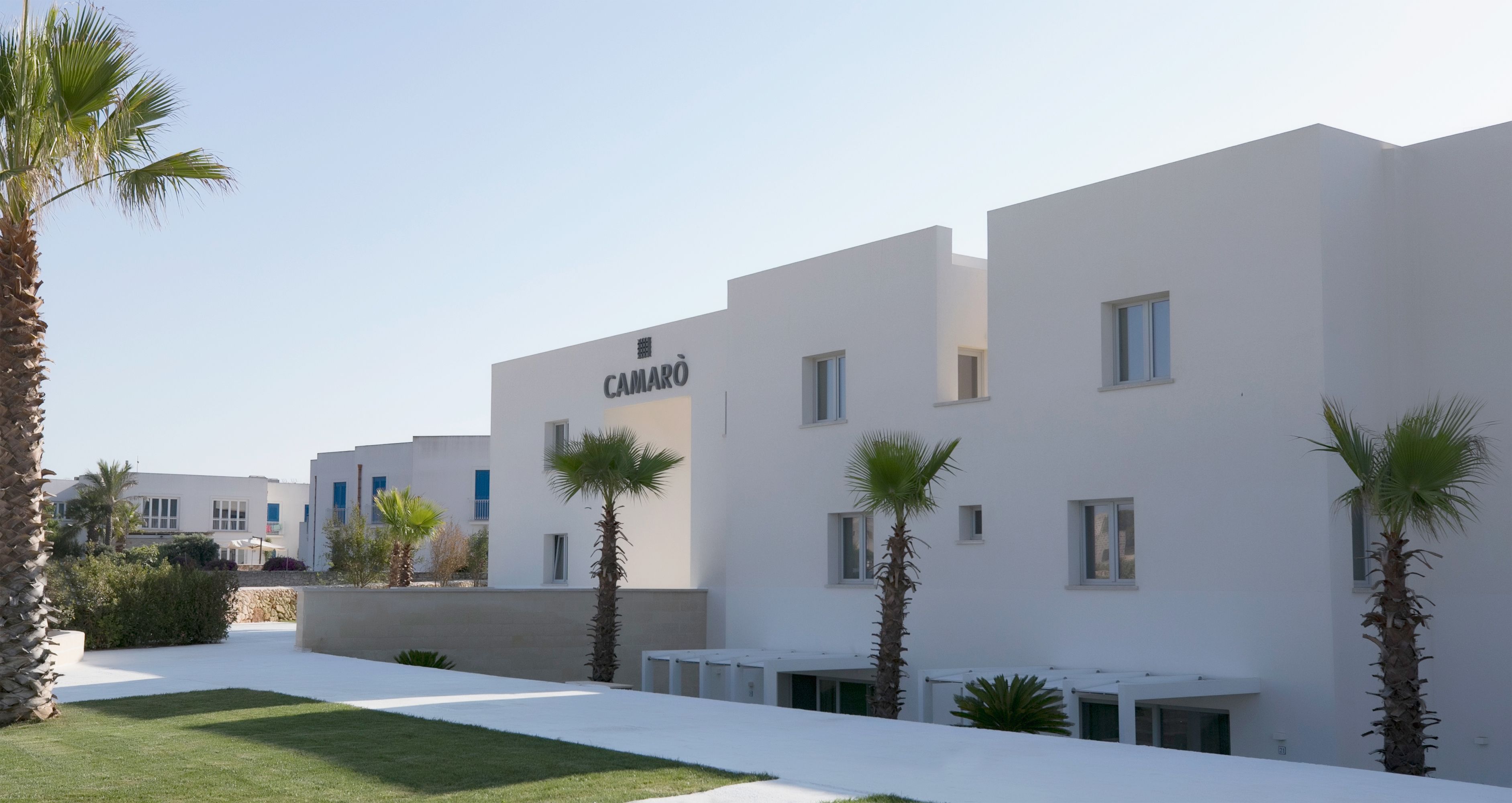 A modern building with the name "CAMARÒ" on the facade, surrounded by palm trees and a well-maintained lawn.