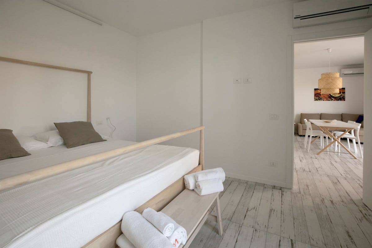 A minimalist hotel room with a single bed, a wooden bench with towels, and a dining area visible through an open door.