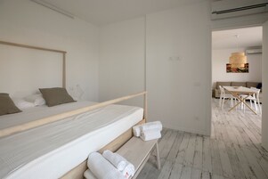 Habitación de hotel minimalista con una cama individual, un banco de madera con toallas y un área de comedor visible a través de una puerta abierta.