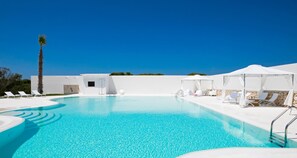 A clear blue swimming pool with white lounge chairs and umbrellas, surrounded by a white building with a palm tree. - Camarò Favignana (Favignana)