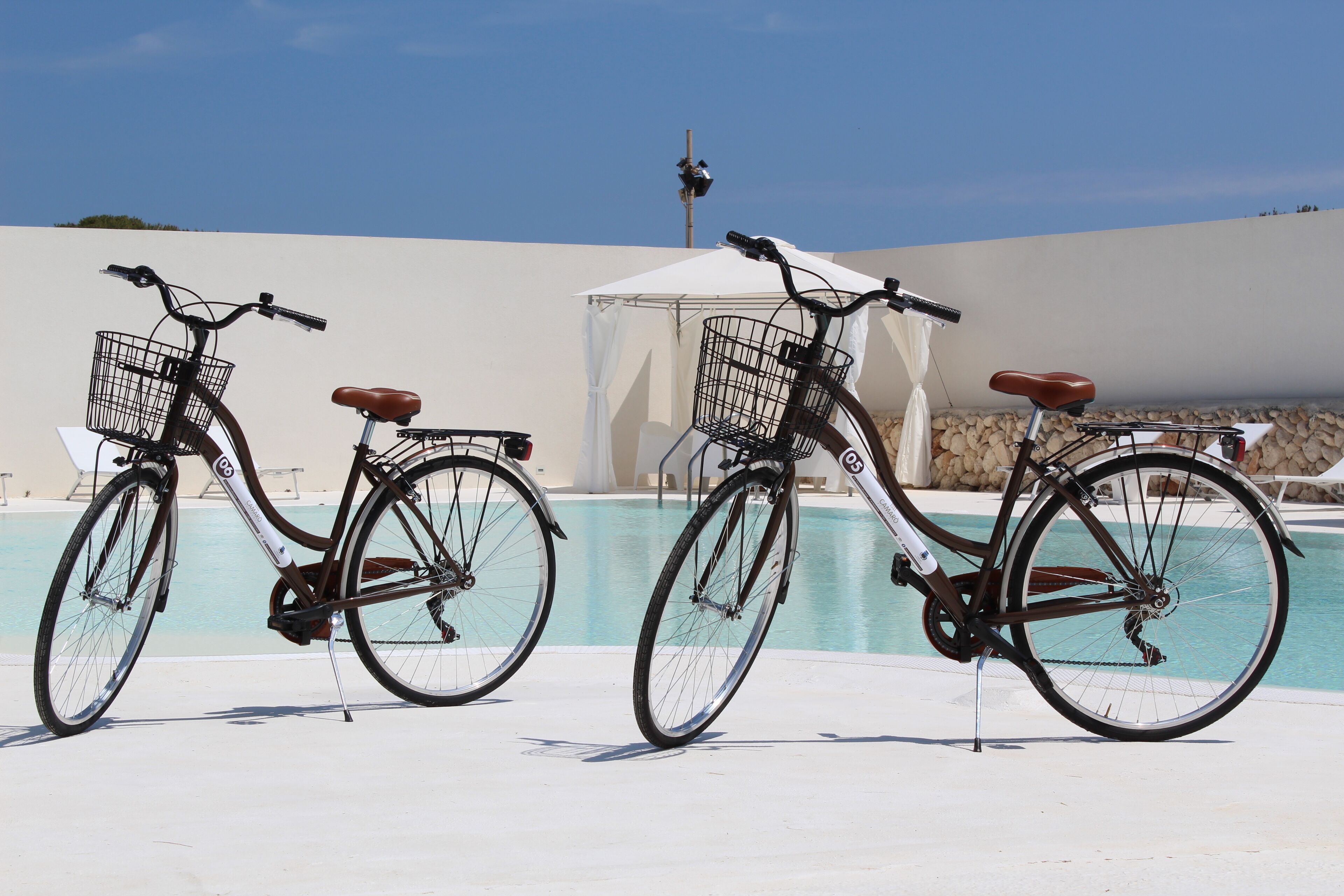 Two bicycles parked by a poolside with a tent and lounge chairs in the background.