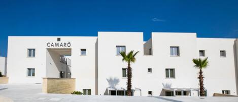 A modern building with the name "CAMARÒ" on the facade, featuring a symmetrical design with multiple windows and palm trees in front.