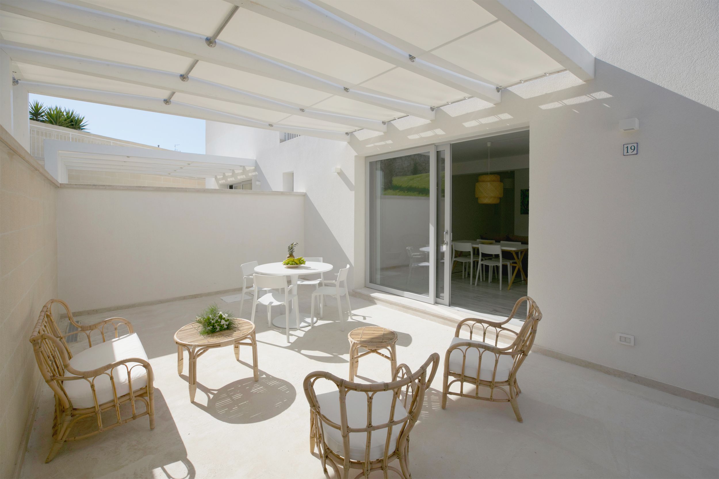 Une terrasse extérieure couverte, aménagée avec des meubles en osier, une table ronde et une porte-fenêtre coulissante donnant sur un espace de salle à manger intérieur.