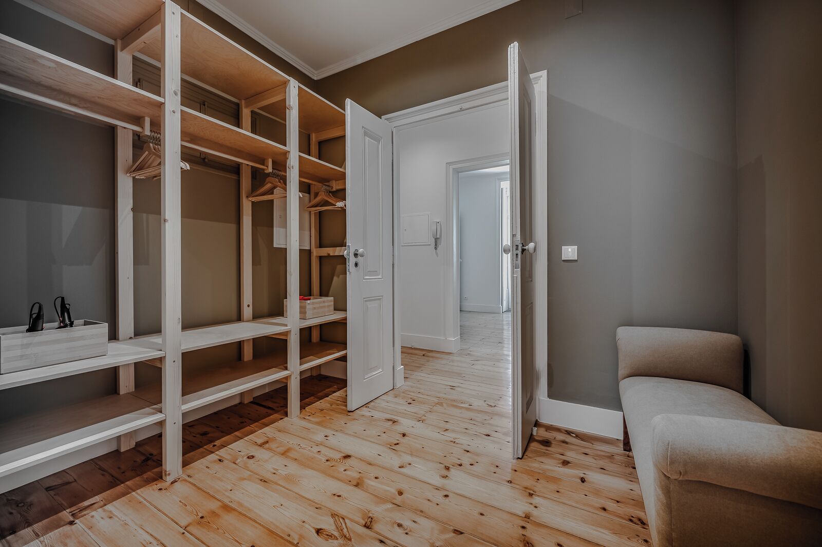 A room with a wooden floor, a white wardrobe with open doors, and a beige armchair.