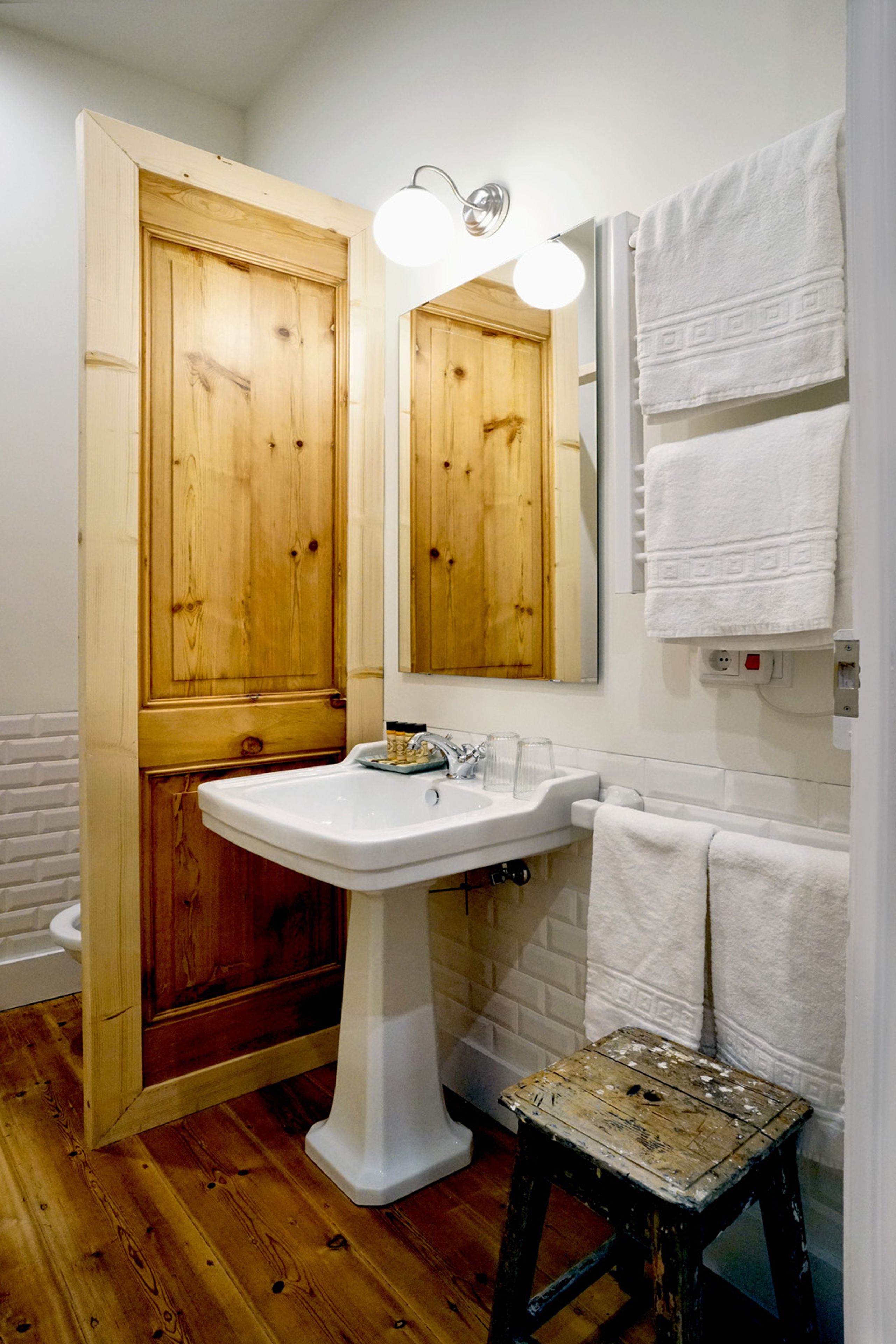 A bathroom with a wooden door, a white sink, a mirror, and a small stool.