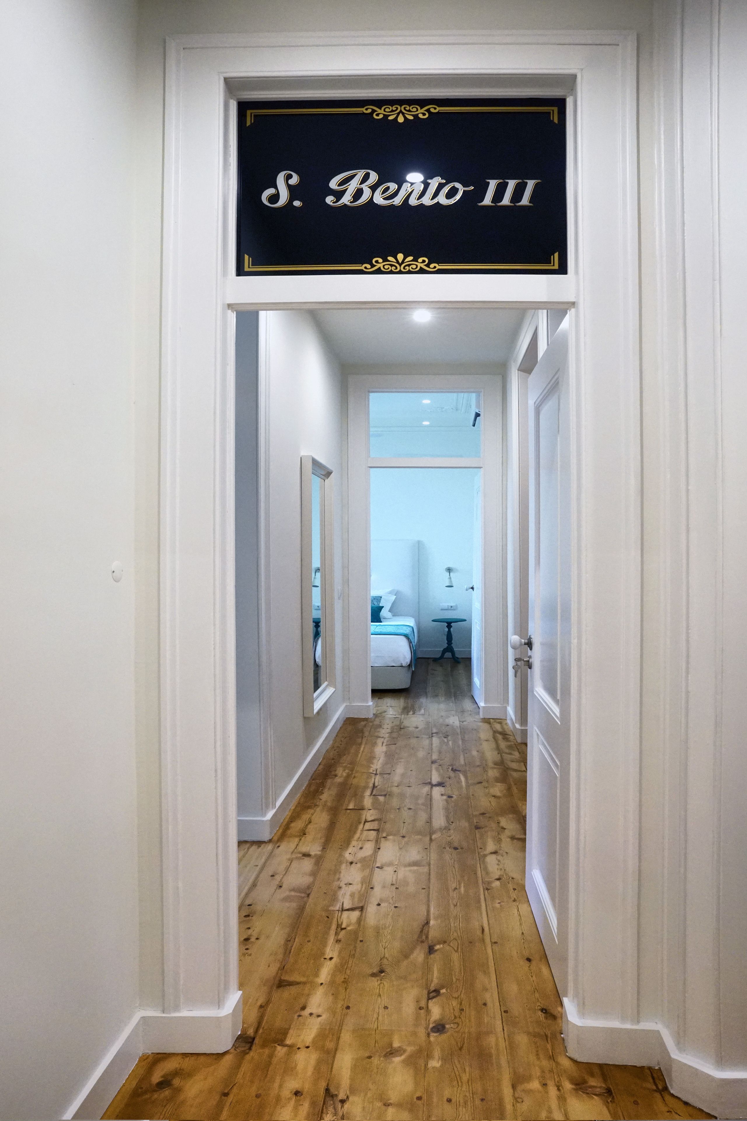 A hallway with a sign reading "S. Bento III" leading to a bedroom with a bed and a nightstand.
