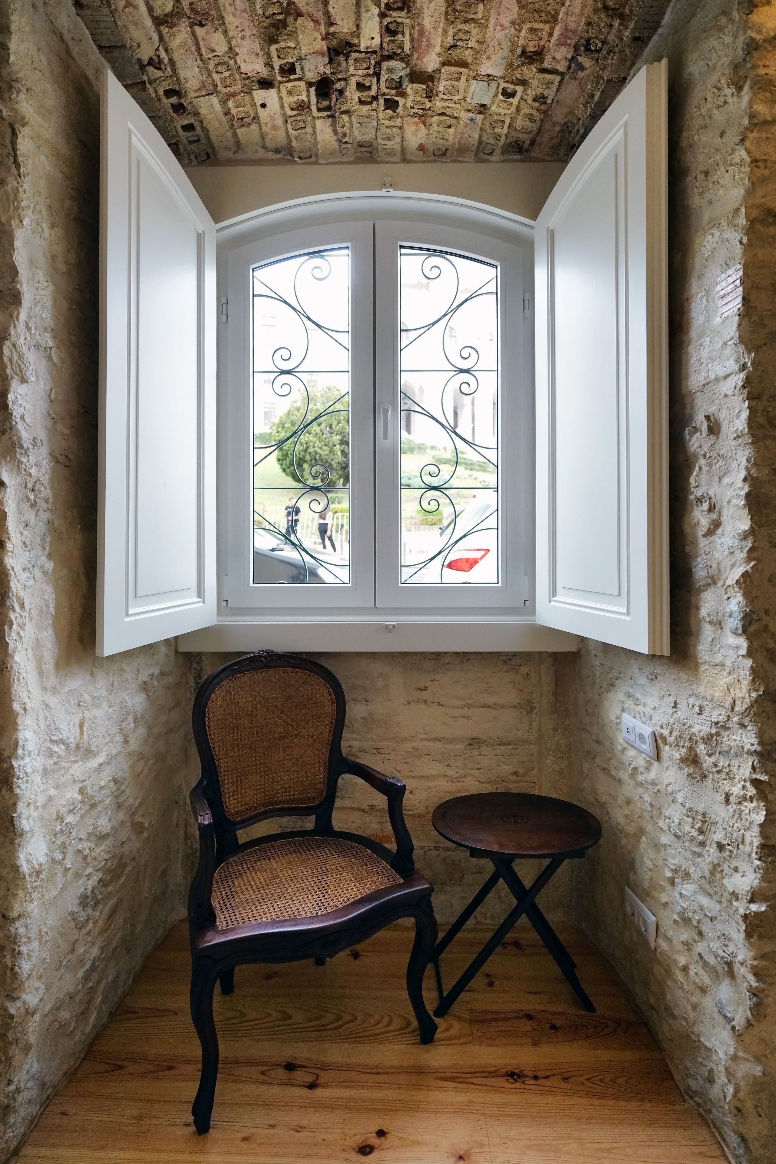A corner room with a rustic ceiling, a window with decorative iron bars, a wooden chair, and a small round table.