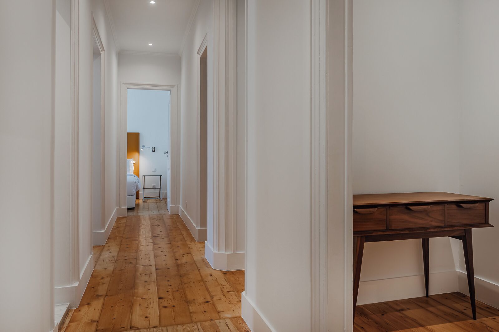 Un pasillo con piso de madera y paredes blancas que conduce a un dormitorio con cama y mesita de noche.