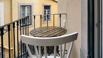 Un balcón con una mesa de madera y sillas blancas, barandal negro y un edificio amarillo al fondo.
