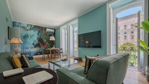 Un salón con un sofá verde, una mesa de centro de vidrio, una zona de comedor con una mesa y sillas, un televisor grande y un mural en la pared.