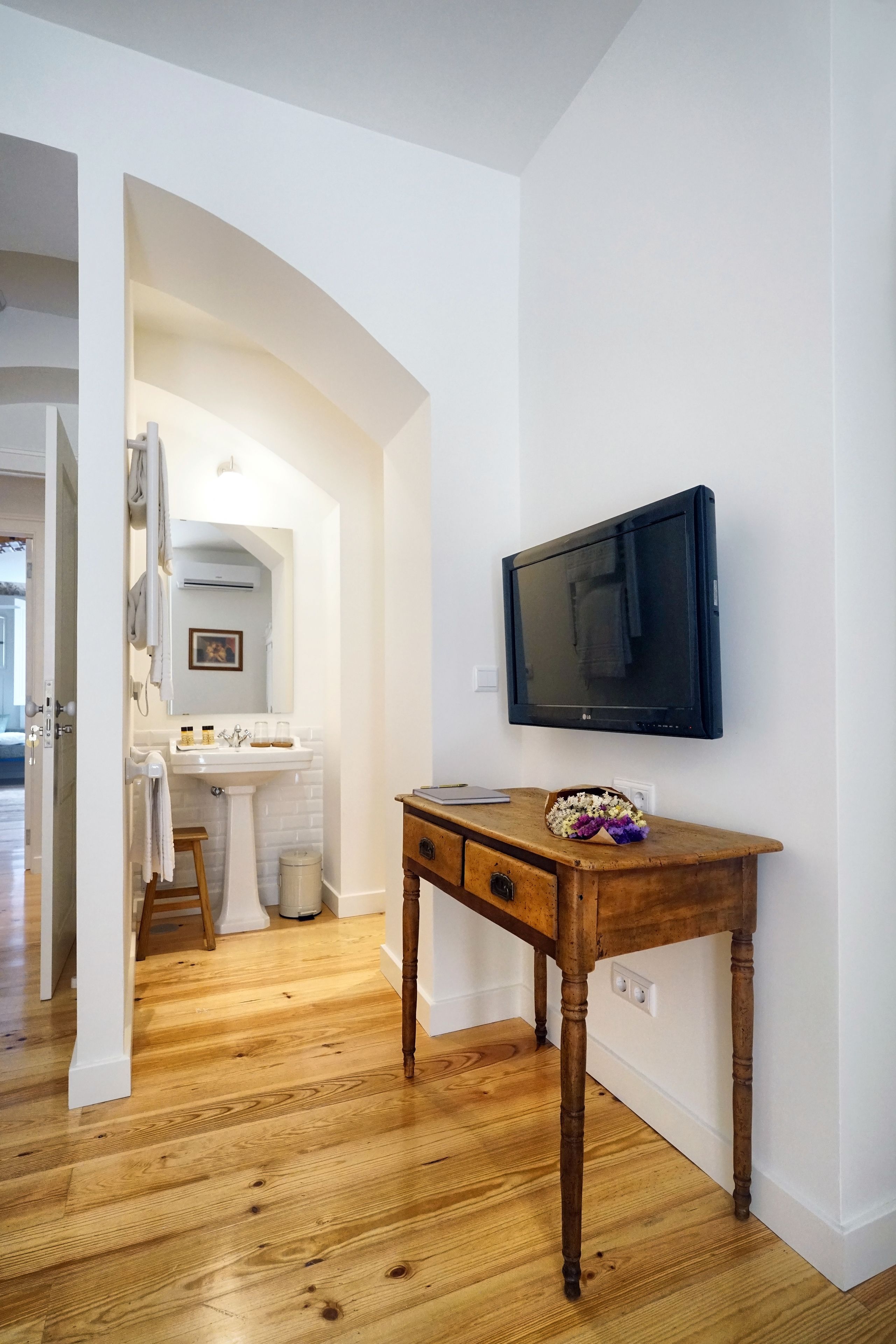 A room with a wooden floor, a white wall, a mounted TV, a wooden table, and a white sink.