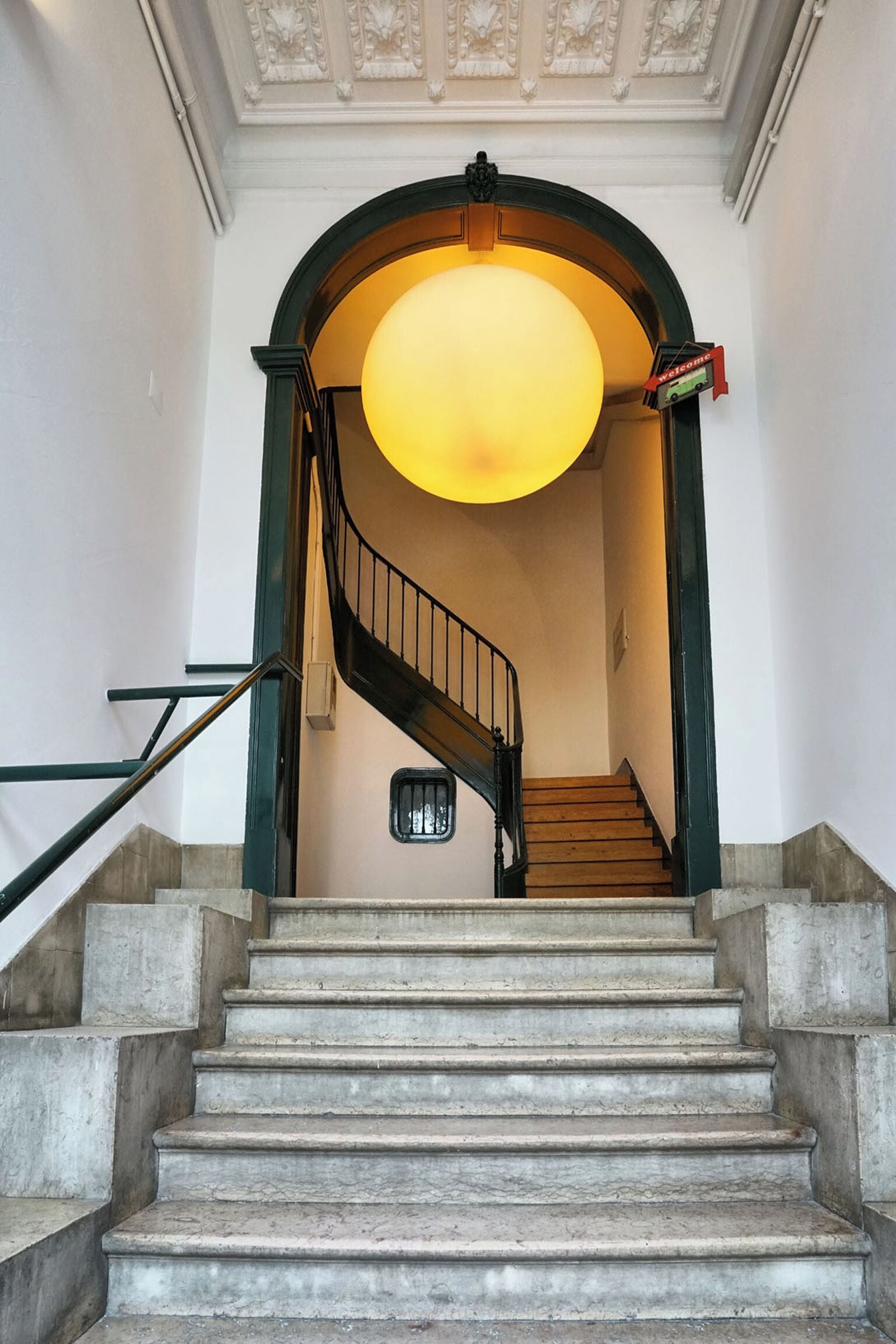 A staircase with a circular light fixture above, leading to a door with a 'Welcome' sign.