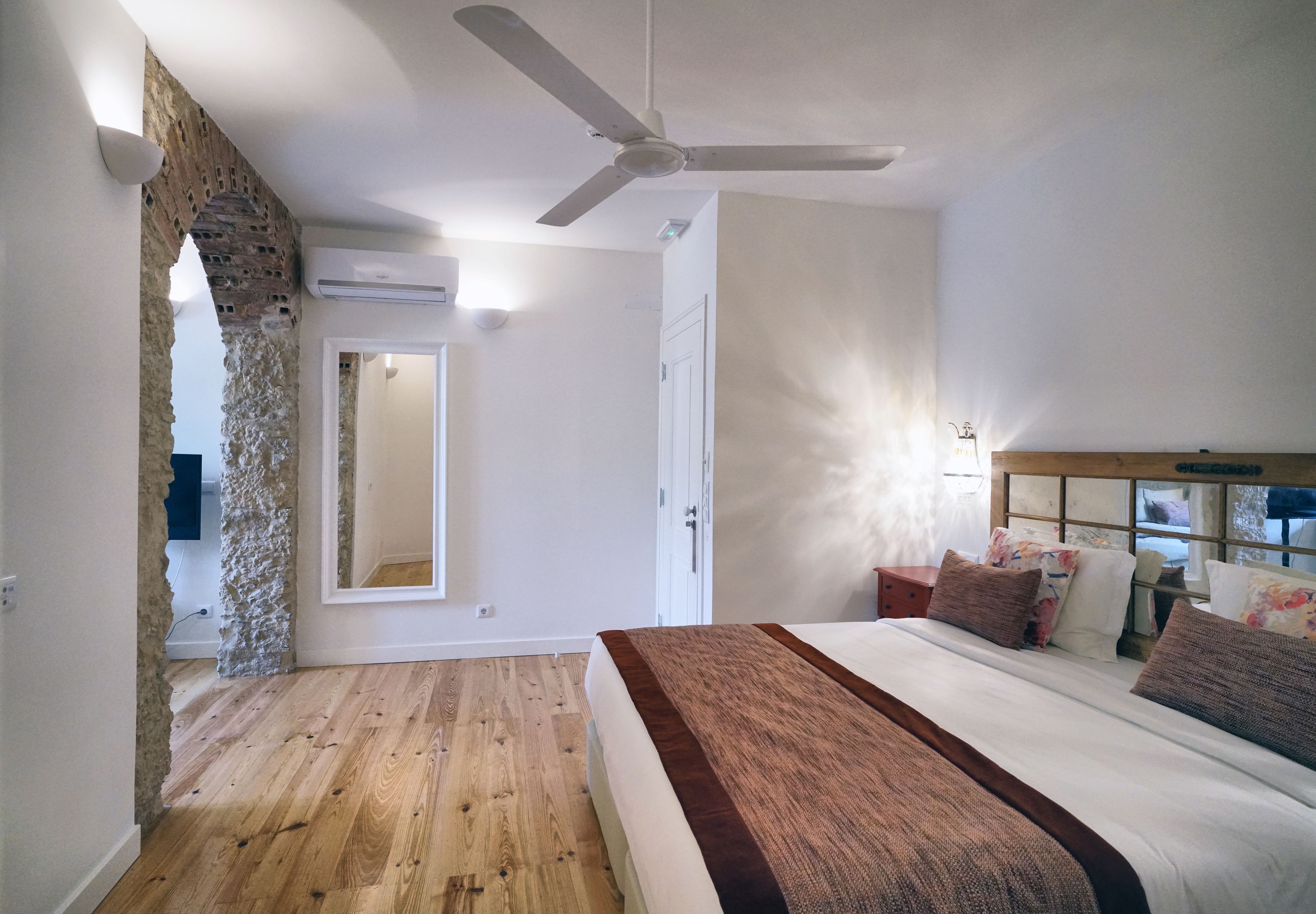 Une chambre avec un plancher en bois, un lit avec une tête de lit marron, un ventilateur de plafond et une arche en briques dans le mur.