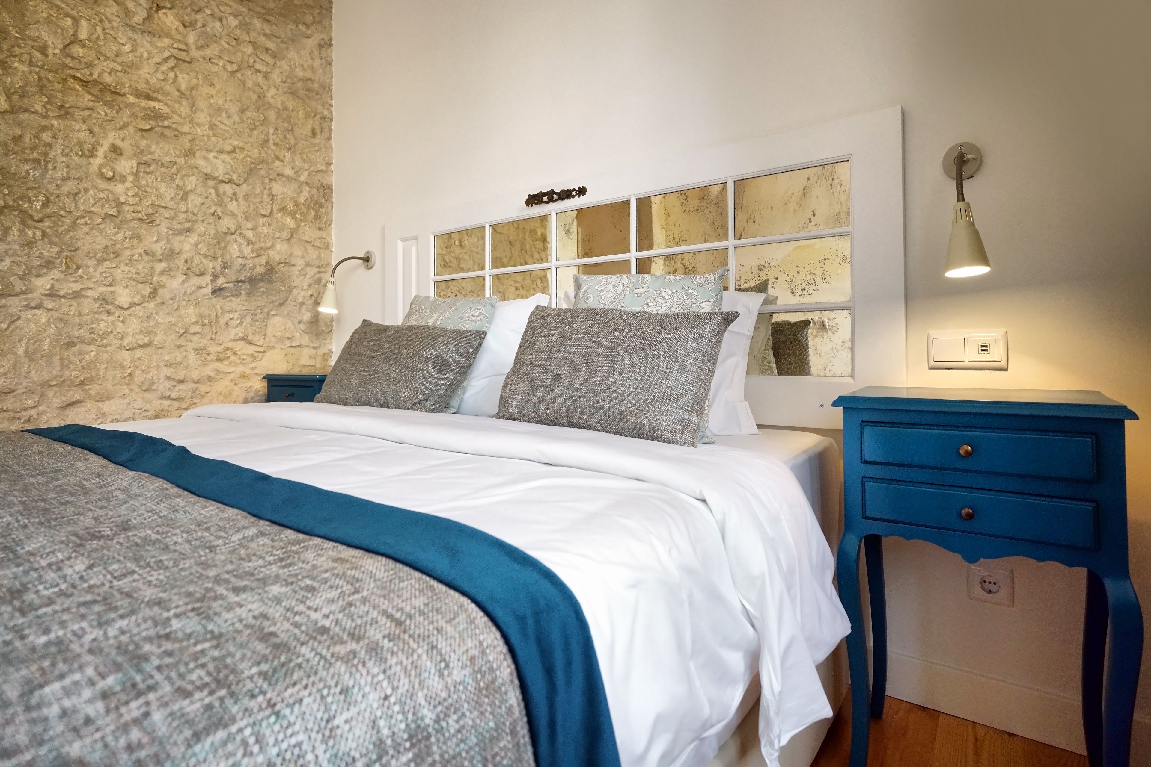A bedroom with a stone wall, a bed with white and gray bedding, a nightstand with a lamp, and a blue dresser.