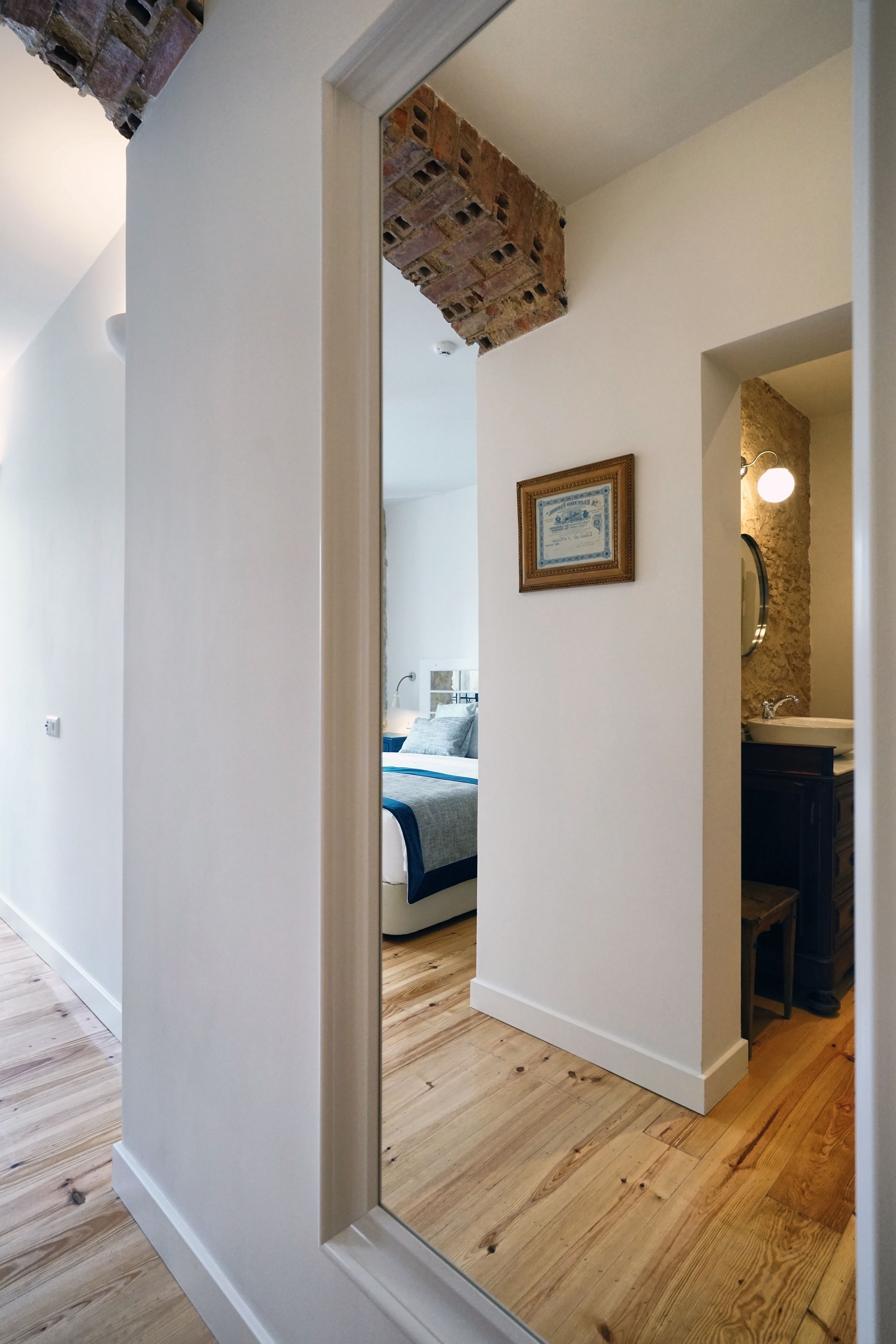 A hallway with a wooden floor, a white wall, and a doorway leading to a bedroom with a bed and a bathroom with a sink and mirror.
