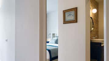 A hallway with a wooden floor, a white wall, and a doorway leading to a bedroom with a bed and a bathroom with a sink and mirror.