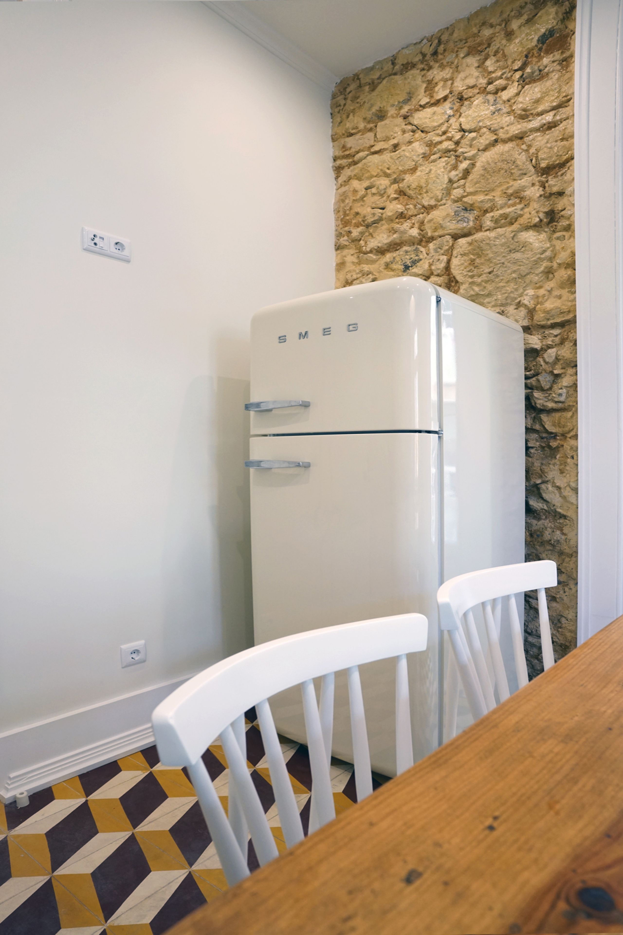 A kitchen with a white refrigerator, white chairs, a wooden table, a stone wall, and a patterned floor.