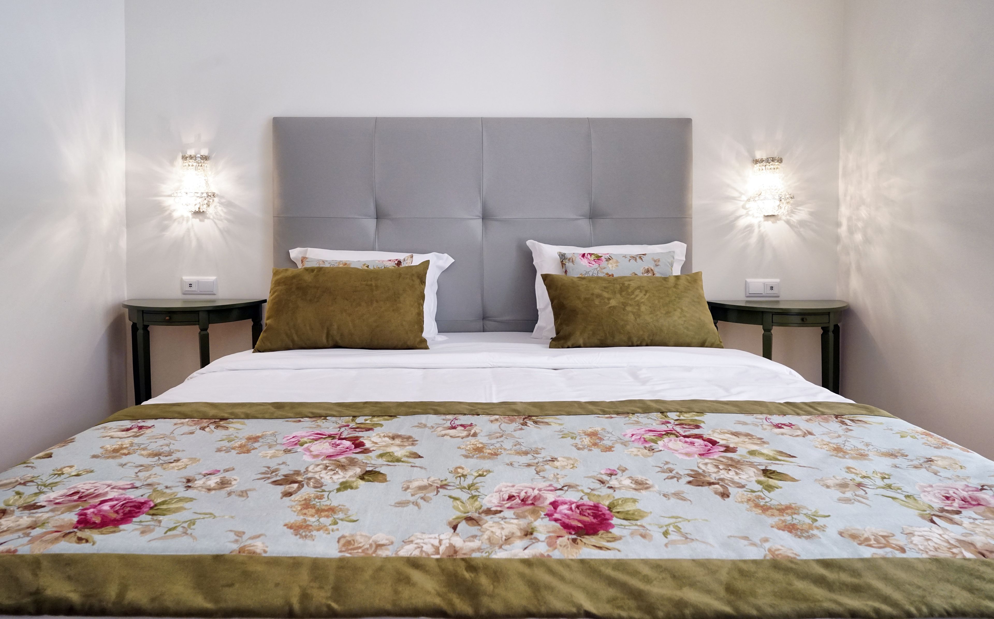 Una cama bien hecha con una colcha floral, dos almohadas y un cabecero tapizado en gris. Hay dos mesitas de noche con lámparas fijadas a la pared.