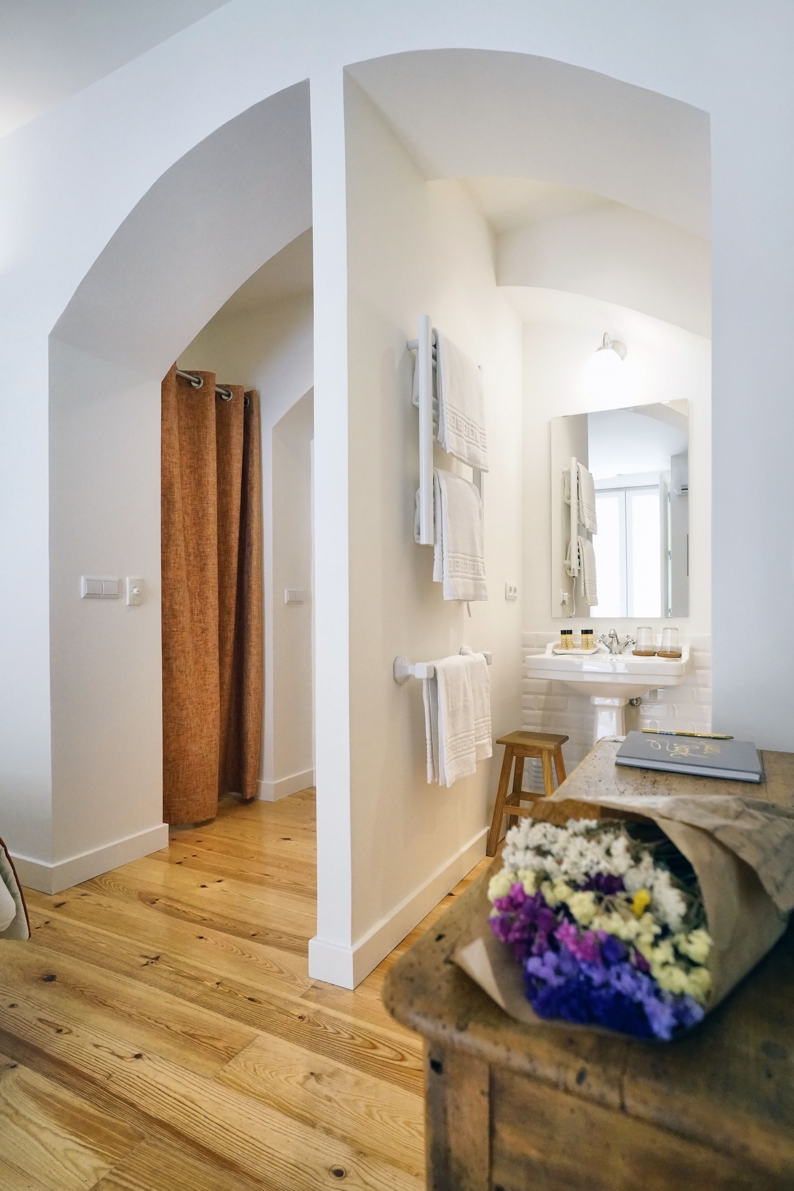 Une salle de bain moderne avec un plancher en bois, des murs blancs, un grand miroir et un tabouret en bois.