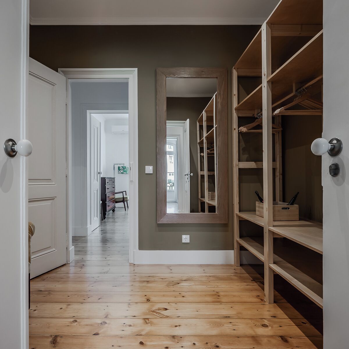 A hallway with wooden flooring, a mirrored closet door, and a view into another room.