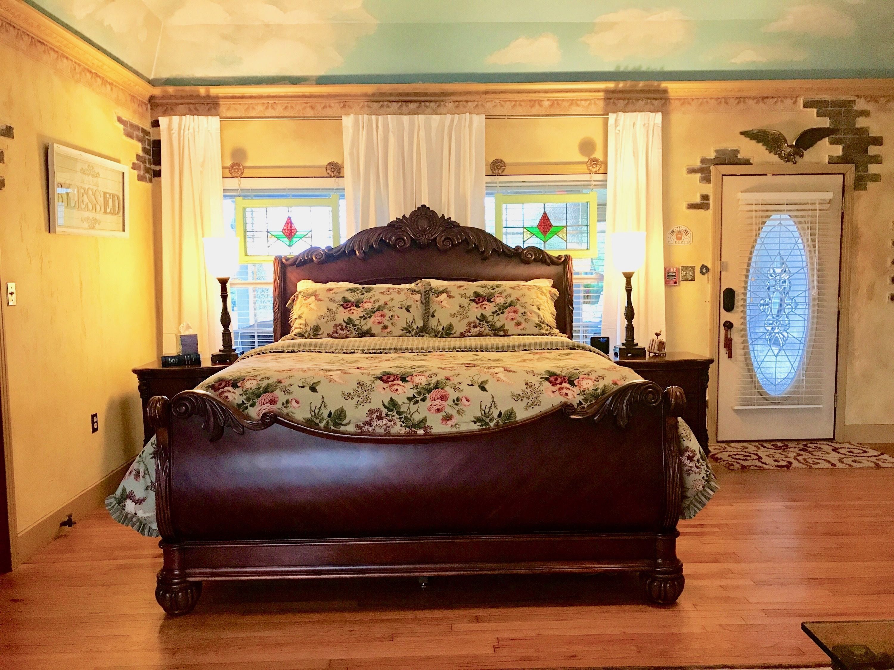 A bedroom with a large bed, a wooden headboard, a nightstand, a lamp, and a window with stained glass.