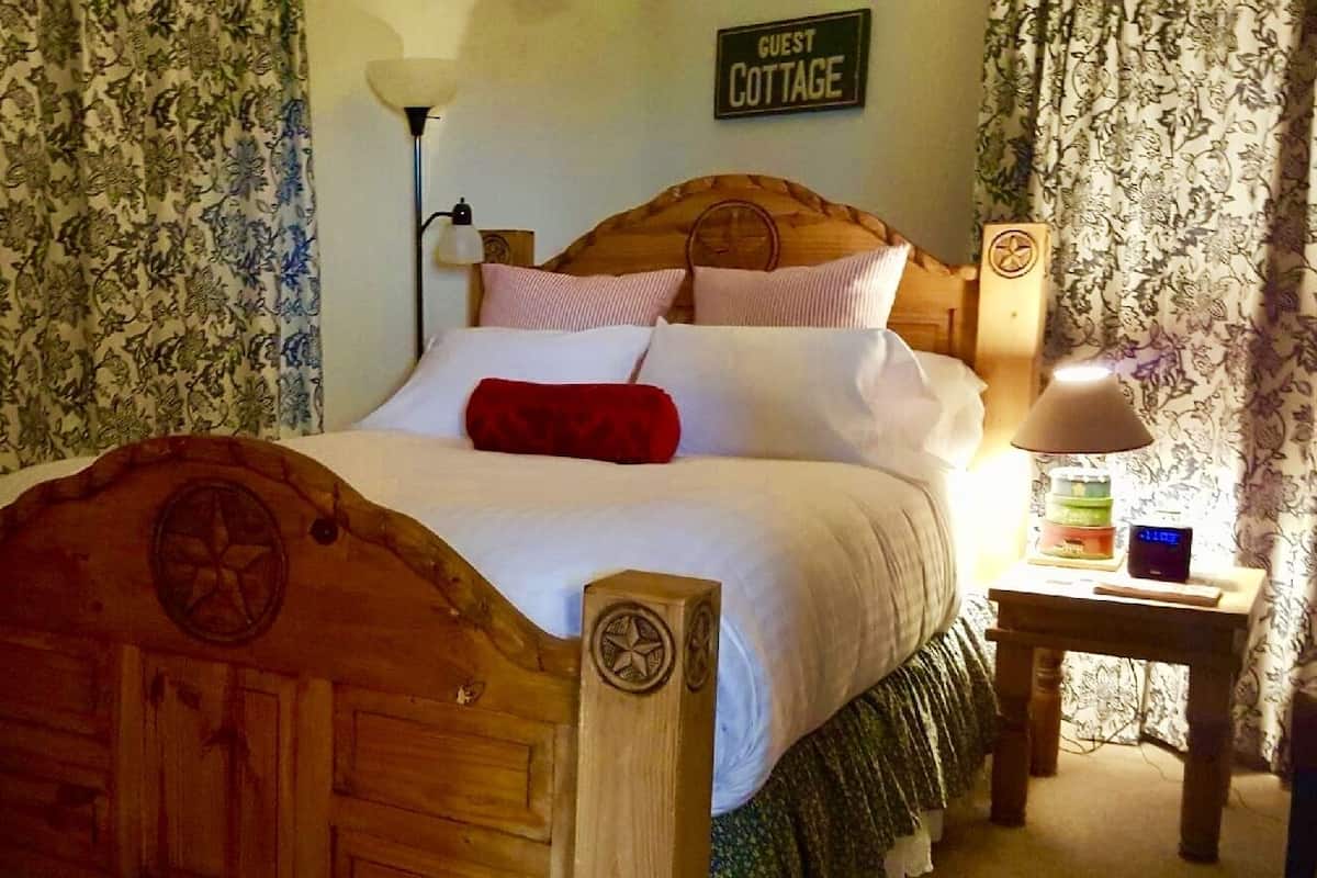 A wooden bed with a headboard, a bedside table with a lamp, and a sign reading "GUEST COTTAGE" on the wall.