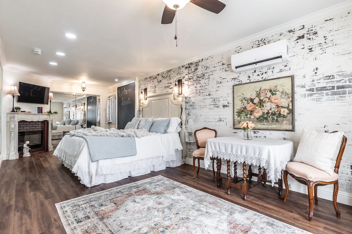 A bedroom with a bed, a table, and a chair, a fireplace, and a brick wall with a painting.